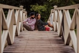 Couple sitting on the bridge back to back, looking at each other near a river at the Painshill Park Surrey engagement shoot
