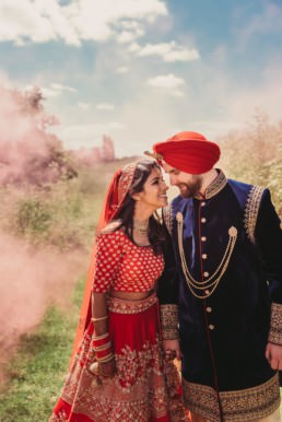 Indiand Bride and Welsh Groom, at a wedding portrait taken in a feild, with smoke grenades added for a pink smoke in the back ground, Bride in red Indina outfit , groom in blue indian outfit, with a turban on